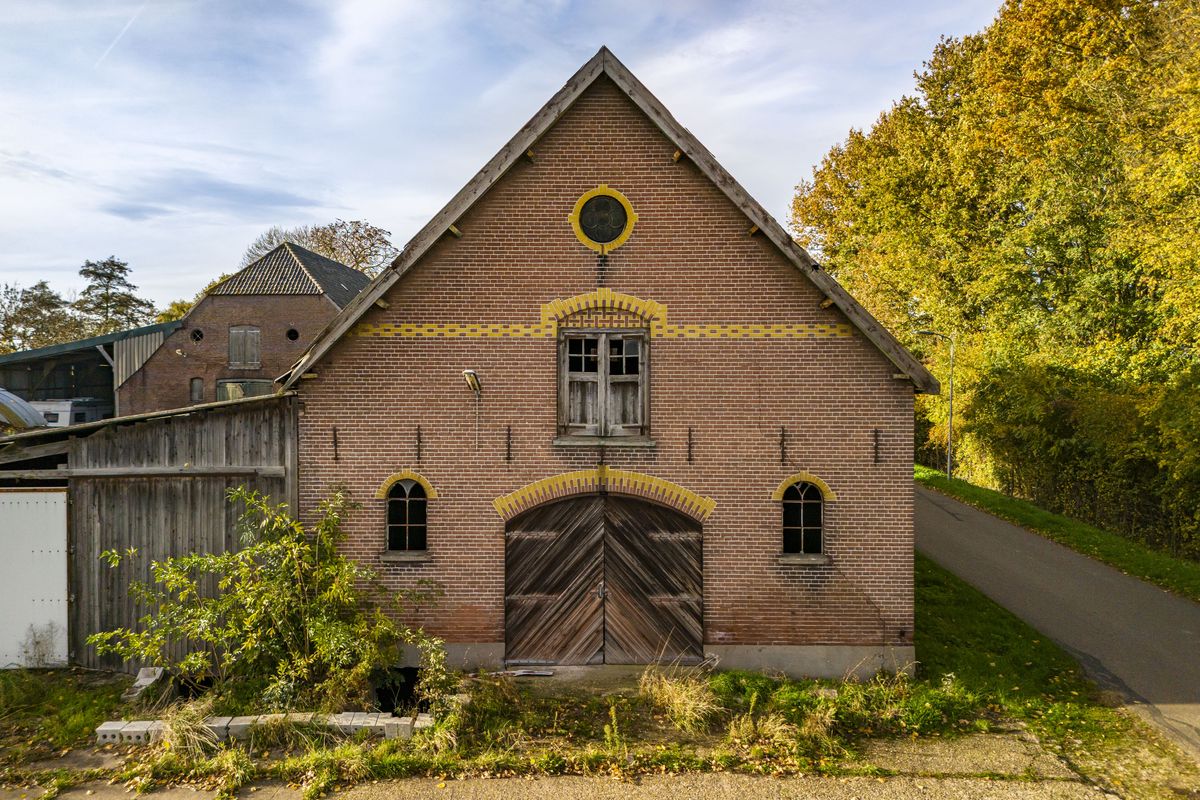 bakstenen schuur met een houten poort, een rond raam en sierlijke banden onder een puntdak in Asch