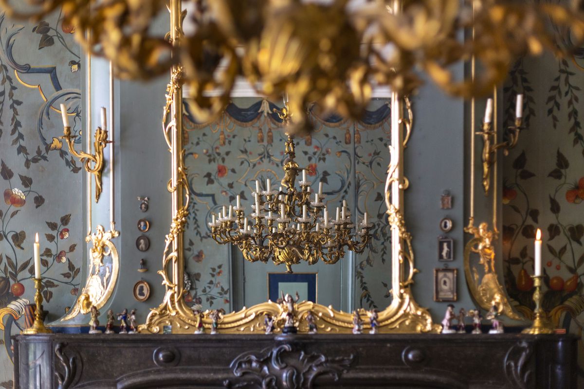 kamer met een gouden kroonluchter, een vergulde spiegel boven een schoorsteen en beschilderd behang met bloemen