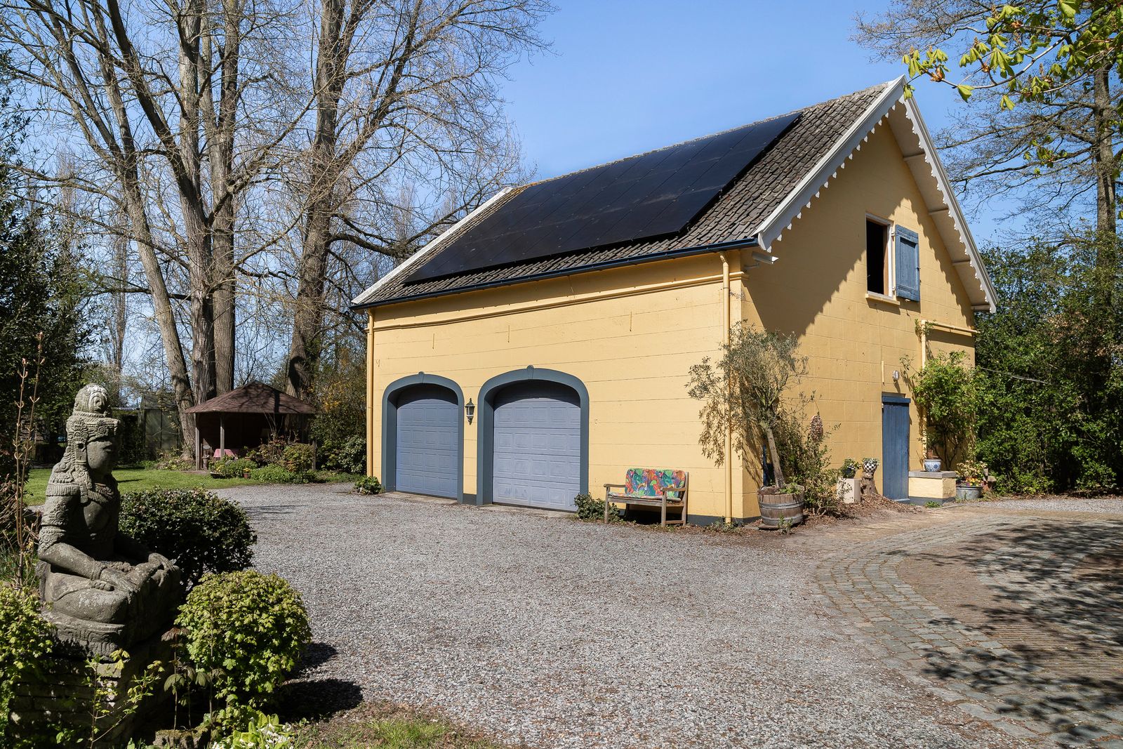 gele garage met zonnepanelen op het dak en twee blauwe garagedeuren tussen hoge bomen in Noordgouwe