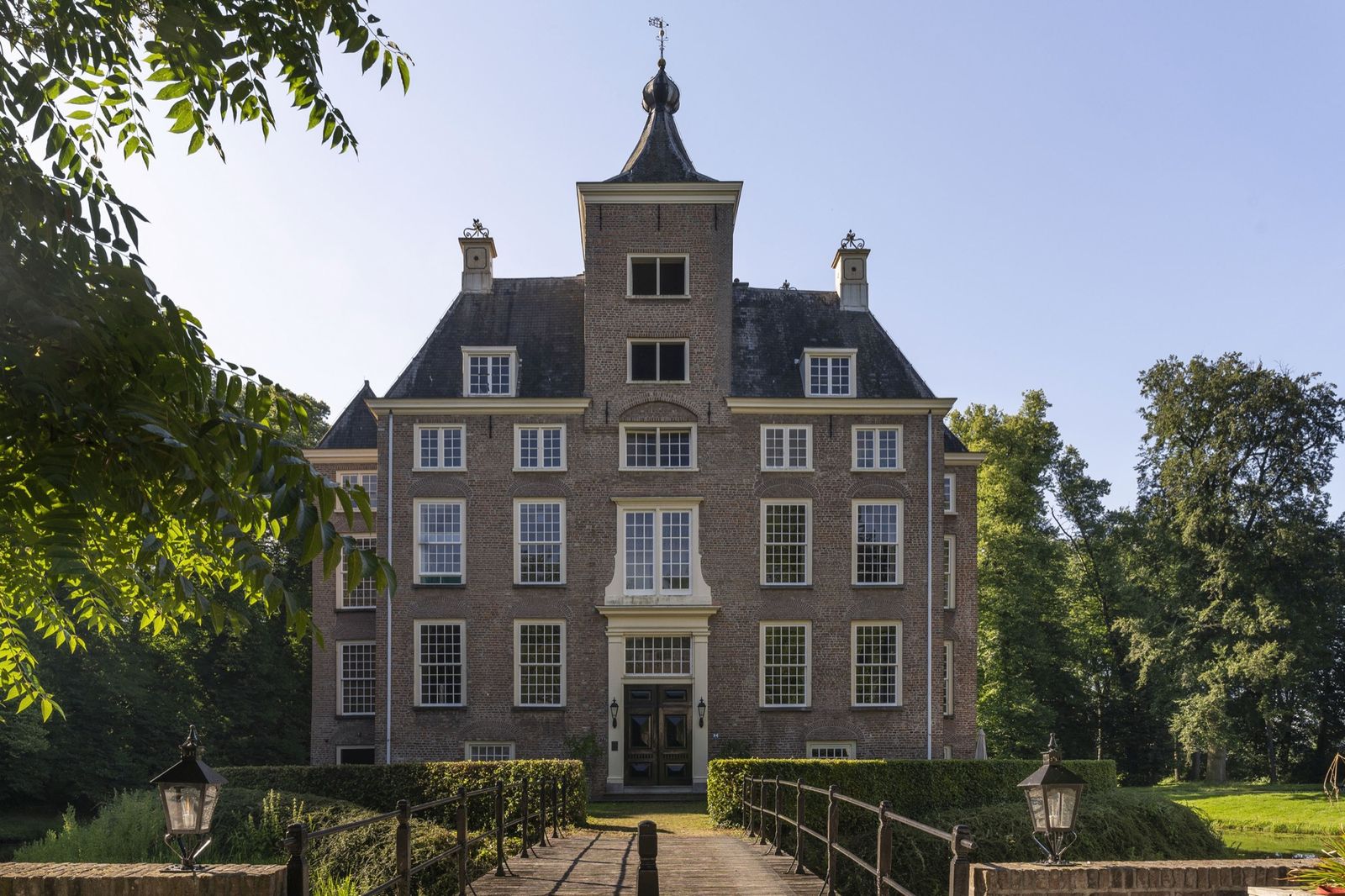 voorgevel van kasteel Soelen in Zoelen, een bakstenen huis met een vierkante toren en een uivormige spits, achter een brug en een haag
